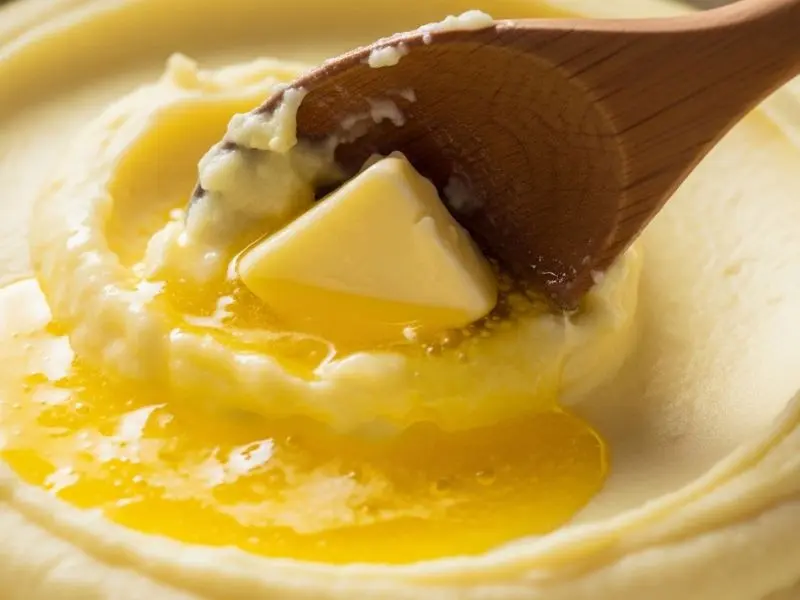 "Purê de batatas sedoso sendo finalizado com manteiga de qualidade amarela intensa criando textura brilhante e cremosa com colher de madeira"