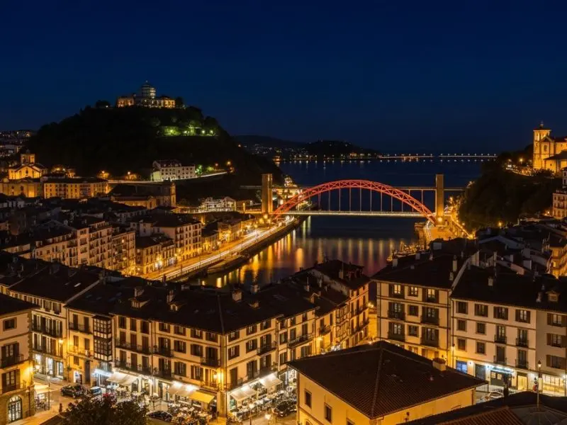 "San Sebastián gastronomia à noite com luzes da Parte Vieja refletindo no rio Urumea e baía ao fundo mostrando cidade viva e culturalmente rica"