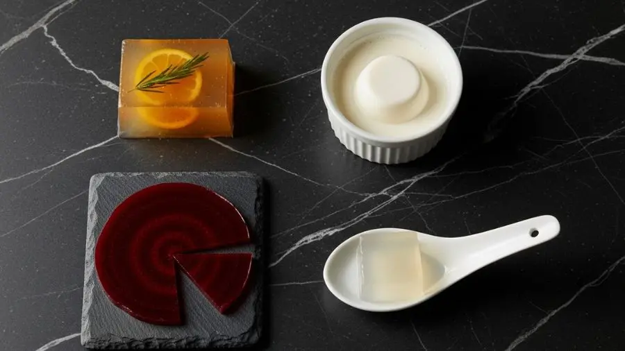 "Arranjo elegante de fotografia de alimentos vista de cima em mármore escuro mostrando quatro preparações distintas de agar-agar como pequenas porções superior esquerdo cubo de aspic de cítrico âmbar-dourado transparente com erva suspensa interior superior direito panna cotta vegana cremosa branca em pequeno ramekin inferior esquerdo lâmina de vidro de beterraba vermelho-profundo translúcido cortada em círculo sobre ardósia escura inferior direito cubo de gel de dashi transparente colocado em colher de porcelana branca mostrando que mantém forma perfeitamente as quatro preparações mostram o alcance das aplicações do agar"