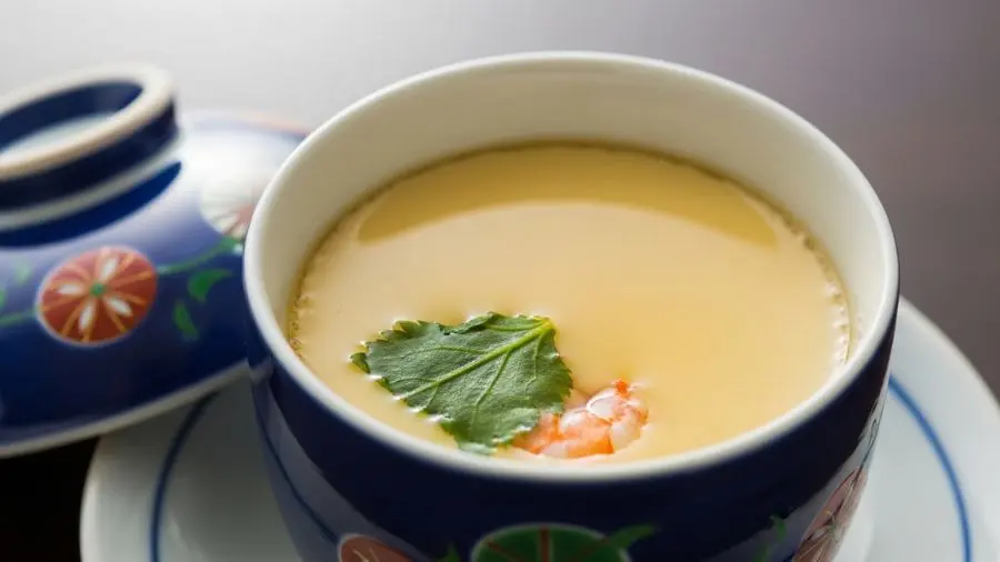 "Close-up de chawanmushi perfeitamente feito em copo de cerâmica japonesa tradicional com tampa ao lado superfície do creme de ovo saboroso perfeitamente lisa e sedosa sem bolhas ou furos indicando cozimento a vapor suave e perfeitamente controlado pequeno pedaço de erva mitsuba e pedaço de camarão apenas visíveis logo abaixo da superfície do creme cor amarelo dourado claro bonito do dashi copo em pequeno prato iluminação lateral natural suave criando brilho suave na superfície do creme cena serena e precisa" 