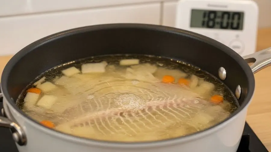 "Close-up do fumet de poisson no estágio frémissement em panela larga e rasa o frémissement mostra apenas o tremor mais suave e delicado movimento quase imperceptível quase como um estremecimento leve nem mesmo bolhas verdadeiras líquido já tem cor dourado muito pálida delicada e clara espinhas de peixe e mirepoix blanc visíveis abaixo da superfície translúcida timer de cozinha no balcão mostrando 18 minutos a cena comunica urgência da janela de 20 minutos sem drama pura precisão"