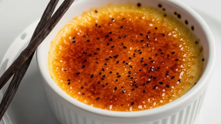 "Close-up lindo de crème brûlée clássico em ramequin de cerâmica branca superfície de açúcar caramelizado perfeitamente uniforme âmbar-dourado profundo mostrando camada de açúcar queimado crocante através do caramelo levemente translúcido o creme de baunilha abaixo visível cor creme com sementes de baunilha pretas claramente visíveis pontos suspensos por toda parte fava de baunilha descansando artisticamente contra o ramequin iluminação dramática de restaurante de cima"