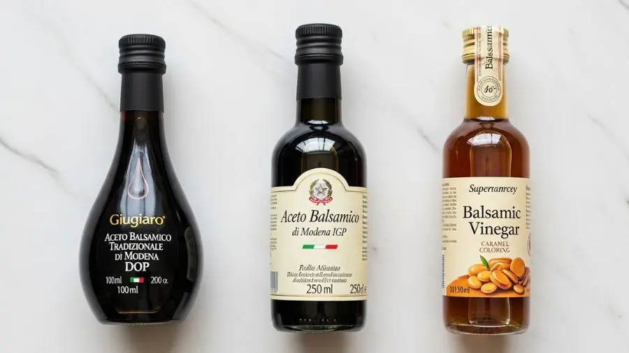"Comparação editorial limpa em mármore branco de três produtos de vinagre balsâmico lado a lado à esquerda garrafa icônica de 100ml formato gota Giugiaro de Aceto Balsamico Tradizionale di Modena DOP escuro pequeno e prestigioso ao centro garrafa média de 250ml de Aceto Balsamico di Modena IGP de produtor italiano de qualidade vidro escuro e rótulo italiano à direita vinagre balsâmico comum de supermercado em garrafa transparente mostrando corante caramel a diferença de qualidade imediatamente clara apenas pela embalagem"