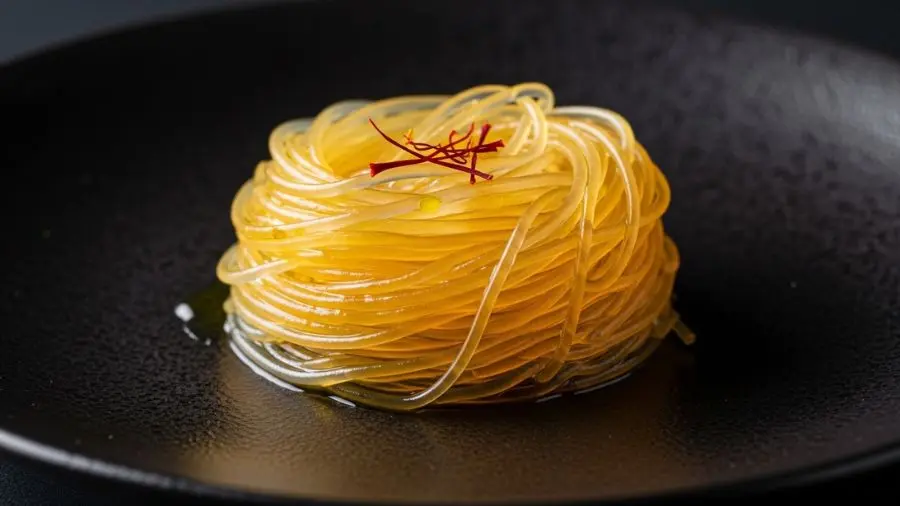 "Foto de arte culinária precisa e bela ninho de espaguete de agar translúcido feito com líquido infusionado de açafrão fios delicados amarelo-laranja semelhantes a vidro dispostos em pequeno ninho compacto em prato de cerâmica escuro os fios são perfeitamente uniformes lisos e brilhantes cada um com cerca de 2mm de diâmetro único fio de açafrão fresco descansando no topo como guarnição pequena quantidade de azeite criando brilho leve o espaguete de gel mantendo sua forma perfeitamente cena elegante moderna inspirada na gastronomia molecular iluminação dramática de restaurante lateral" 