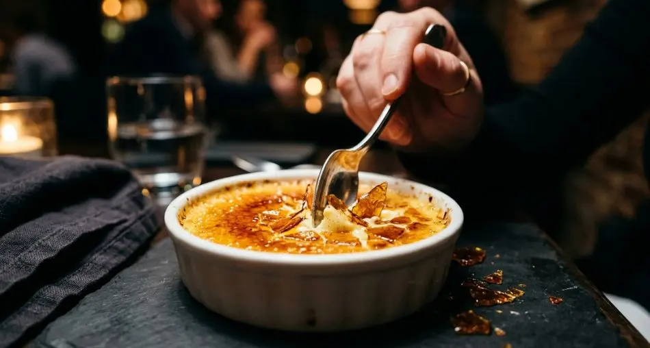 Perfect crème brûlée in white ramekin, golden amber caramel crust being cracked by a silver spoon, dark moody background, dramatic side lighting, fine dining photography, 4k